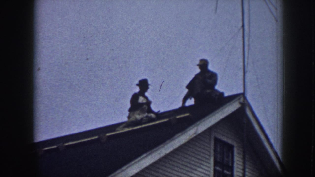 Two silhouetted male figures on a clapboard house roofline, Nebraska 1958, with a thin overhead wire visible, captured on 8mm home-movie film.