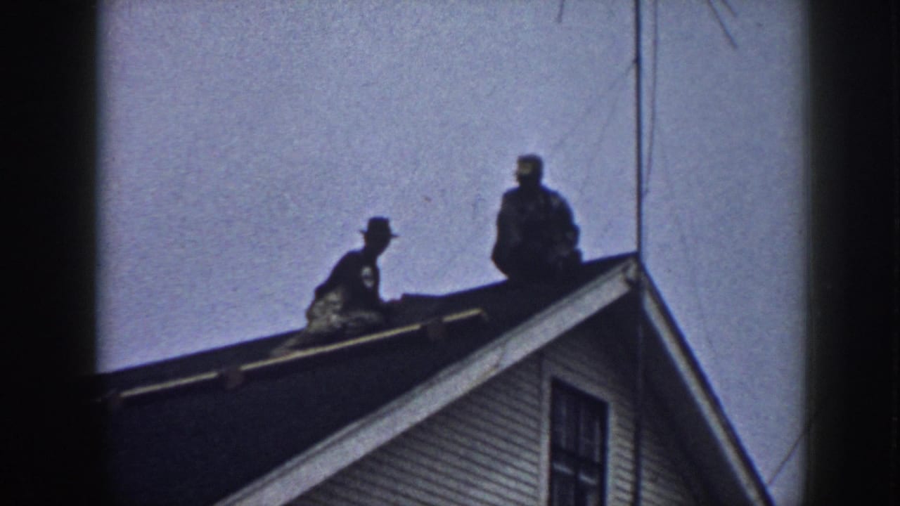 Two men in work hats on the ridge of a clapboard-sided house, Nebraska circa 1958, silhouetted against a pale sky on 8mm film.