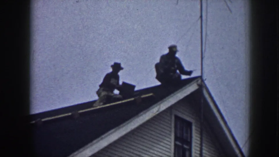 Two men working on a clapboard house rooftop, Nebraska 1958, one holding a rectangular object, silhouetted in 8mm home-movie footage.