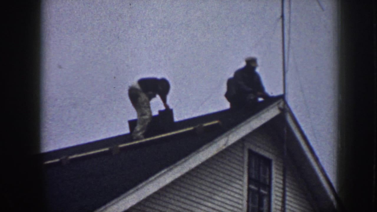 Two men silhouetted on a residential rooftop, one bent near the ridge, the other standing; clapboard house exterior and a window visible below.