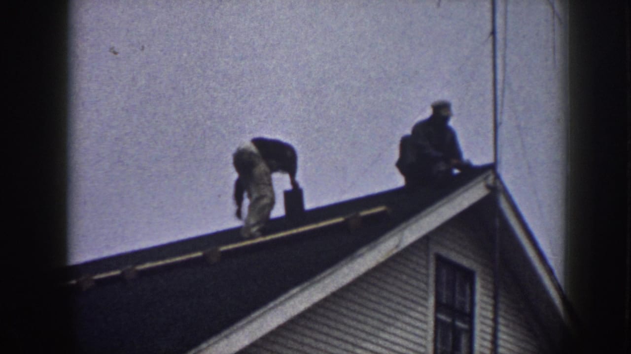 Two silhouetted men on a pitched roof, one crouching with a dark object; clapboard house siding and a window visible below.