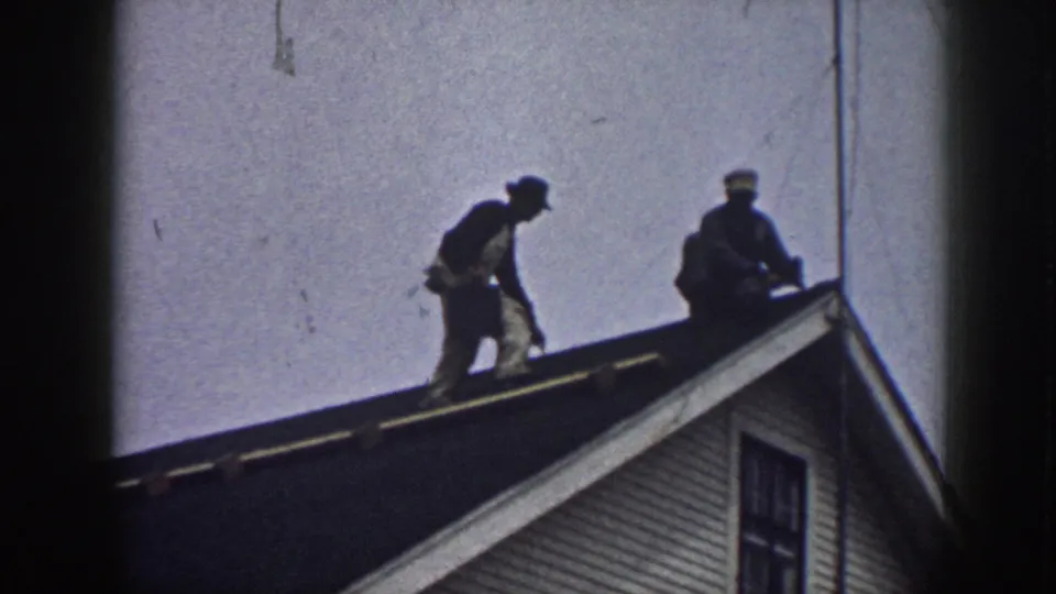 Two male workers on a gabled roof circa 1958, one stooped and carrying what may be roofing supplies, shot from ground level against a pale sky.