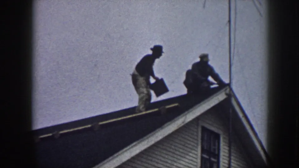 Two men on a residential rooftop circa 1958, one upright with a dark container, the other bent working alongside, white clapboard siding below.