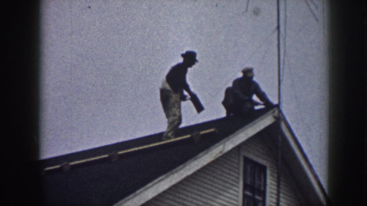 Two men silhouetted working on a residential rooftop in what appears to be Nebraska, 1958; white clapboard siding on the gable below.