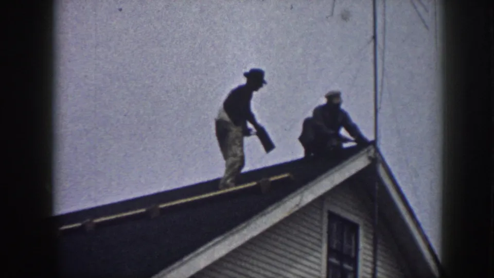 Two men silhouetted working on a residential rooftop in what appears to be Nebraska, 1958; white clapboard siding on the gable below.