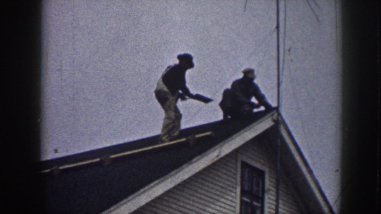 Two men on a house roof peak; standing figure holds an object while the other crouches beside him.