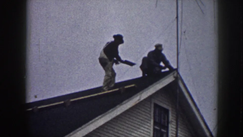 Two men working at a residential roof peak, circa 1958; clapboard siding and a gable window visible beneath them.