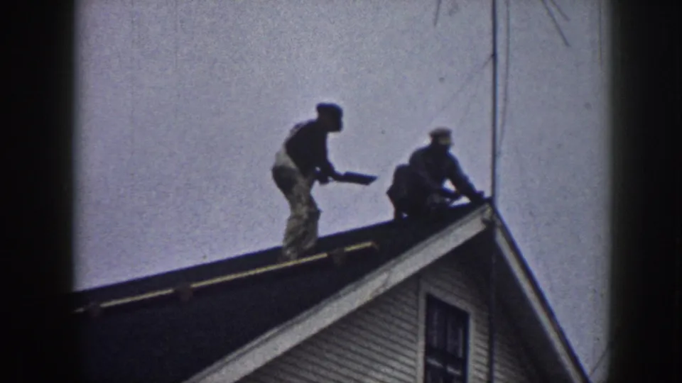 Two men mid-action on a residential rooftop consistent with 1950s Nebraska footage; clapboard siding and window visible below.