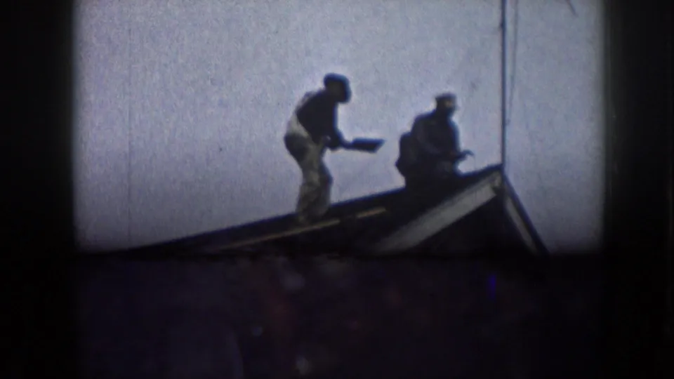 Two men positioned along a house roofline, silhouetted against a pale overcast sky, in footage consistent with late 1950s Nebraska.