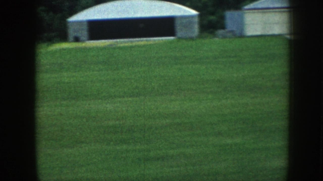 8mm home movie ground shot of a small rural airfield in Nebraska, c.1958, showing a mowed grass strip and metal-clad hangars against a tree line.