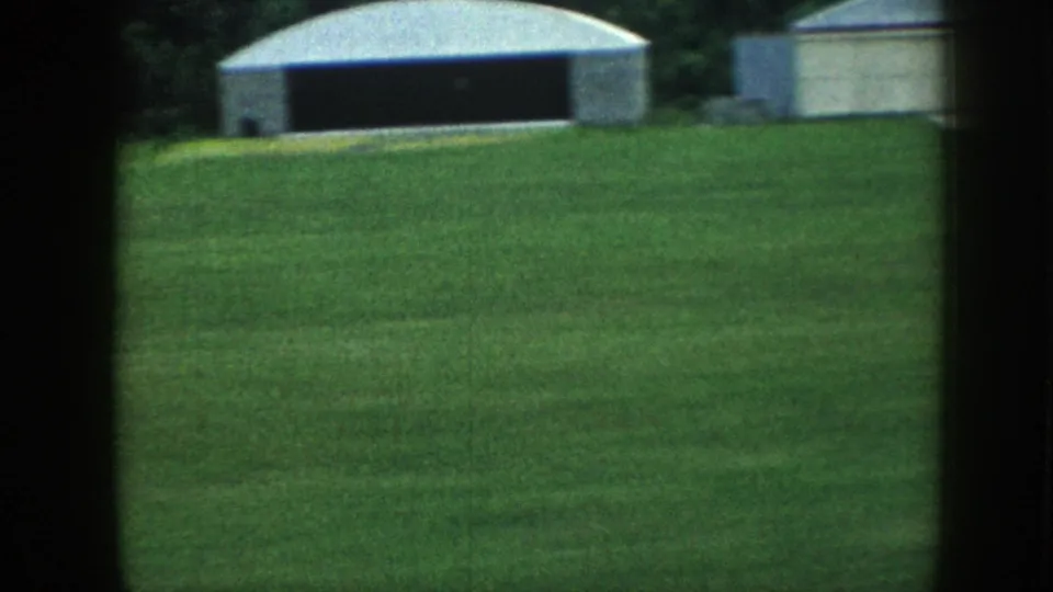 8mm home movie ground shot of a small rural airfield in Nebraska, c.1958, showing a mowed grass strip and metal-clad hangars against a tree line.