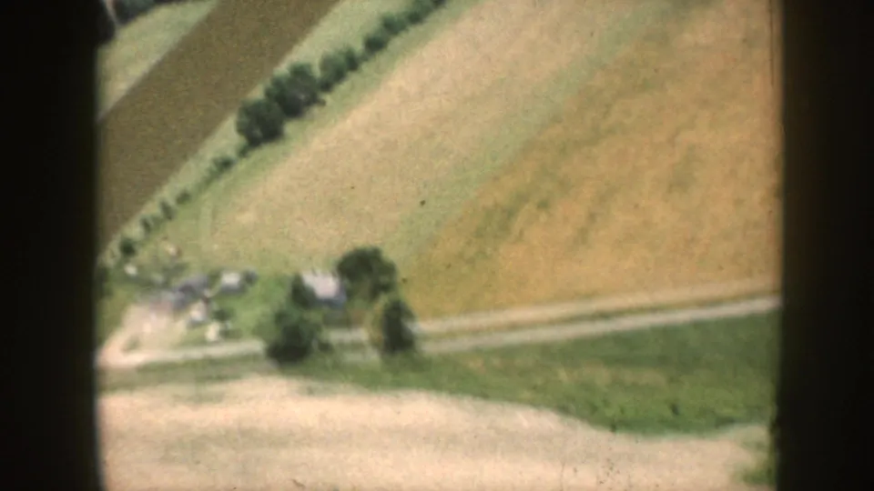 Airborne 8mm shot from above a Midwestern agricultural landscape, c.1958, showing strip-farmed fields, shelterbelts, and small farm buildings below.