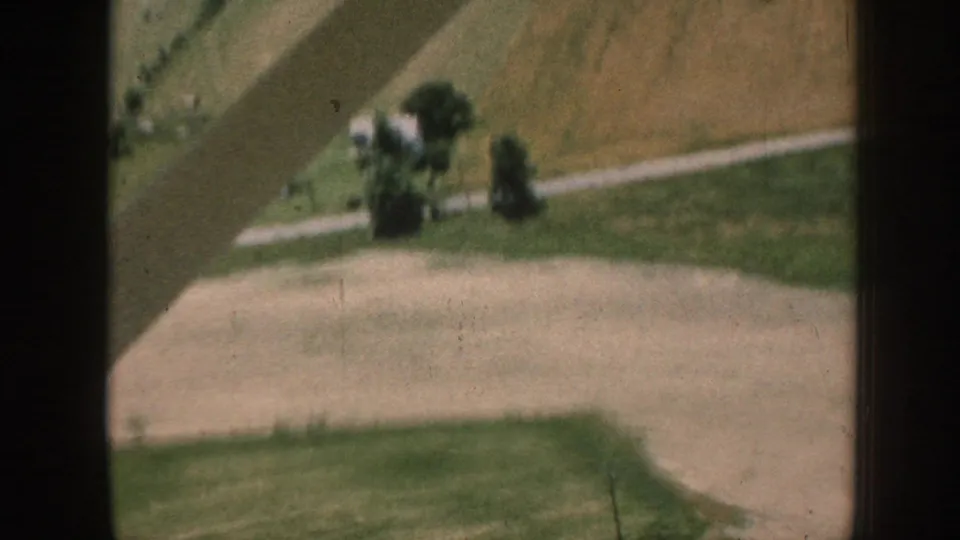 8mm aerial view from a climbing light aircraft over rural Nebraska, c.1958, showing patchwork farmland, tree rows, and a road or track below.