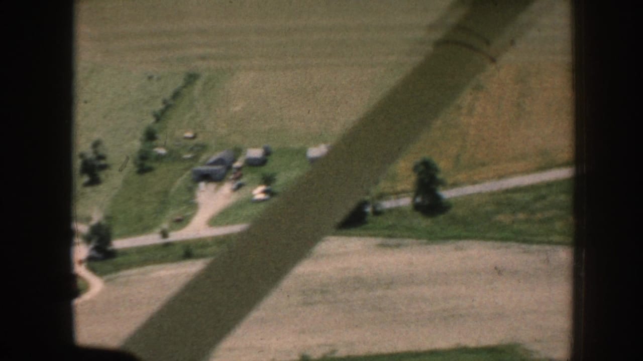Aerial 8mm home movie, c.1958 Nebraska, showing green and golden crop fields, a farmstead cluster with outbuildings, gravel roads, and a diagonal aircraft wing shadow.