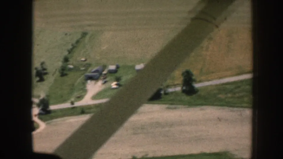 Aerial 8mm home movie, c.1958 Nebraska, showing green and golden crop fields, a farmstead cluster with outbuildings, gravel roads, and a diagonal aircraft wing shadow.