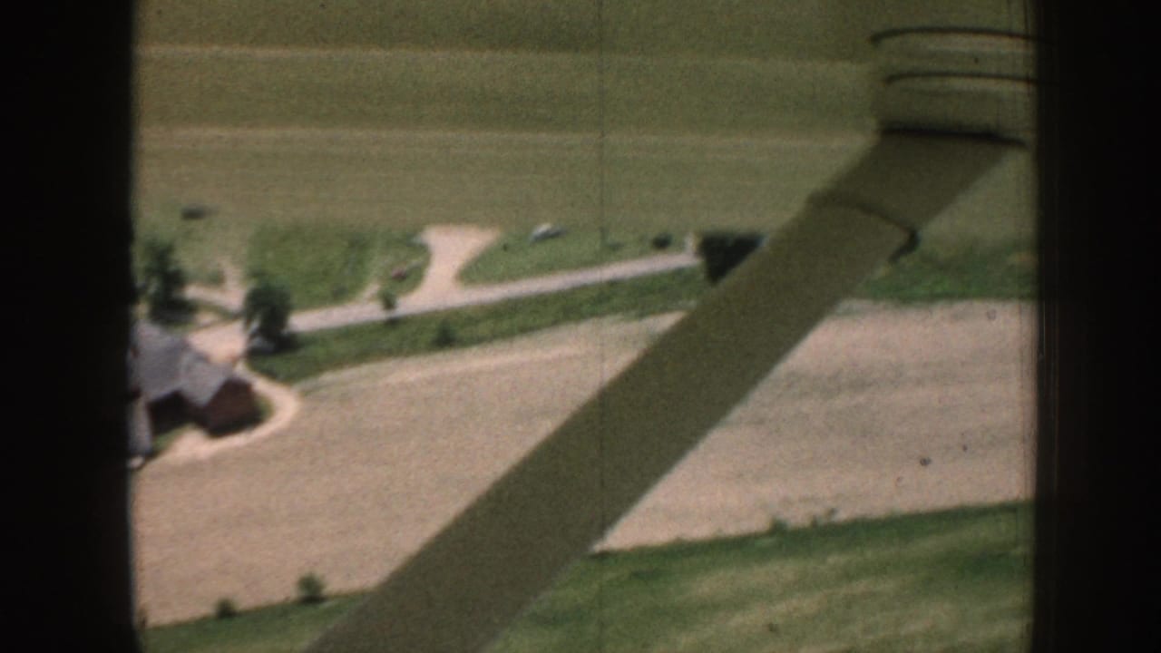 Aerial 8mm frame showing a red barn and white farmhouse amid fields, with a diagonal wing shadow across the landscape.