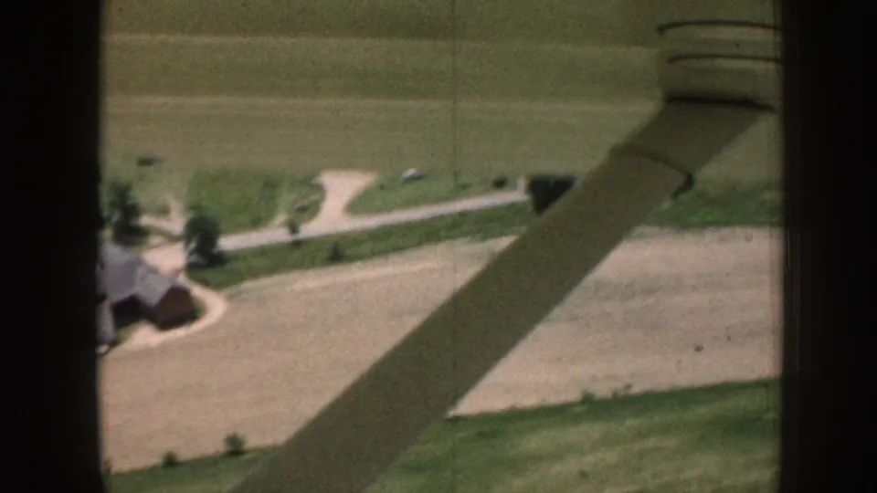 Aerial 8mm home movie, c.1958 Nebraska, showing a red barn, white farmhouse, gravel driveway, and surrounding crop fields under an aircraft wing shadow.