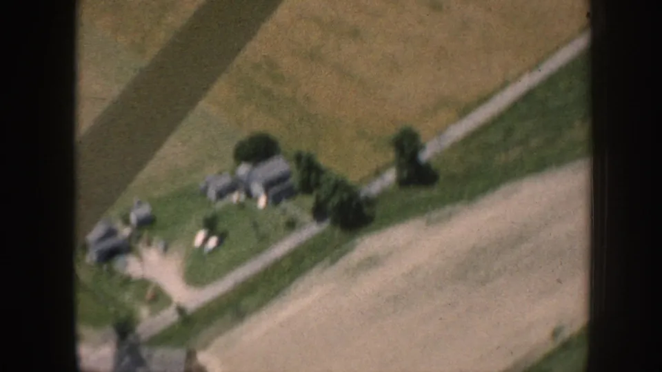 Aerial 8mm home movie, c.1958 Nebraska, showing two farm homesteads with outbuildings, shade trees, white livestock in yards, and a curving gravel road.