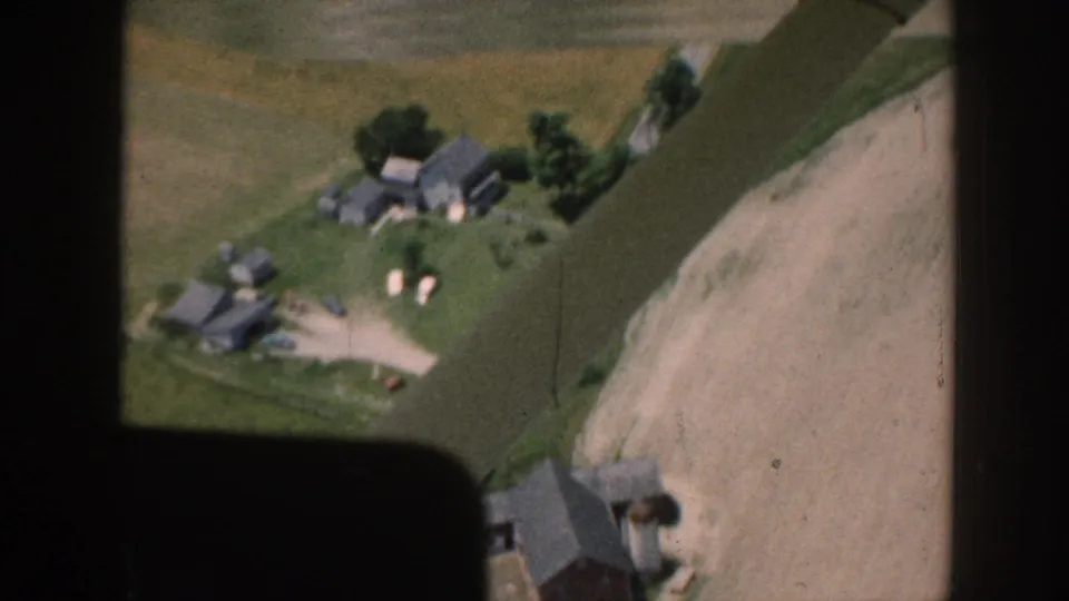 Aerial 8mm home movie, c.1958 Nebraska, showing multiple farm outbuildings, shade trees, white livestock, and a pale sandy field, with the aircraft edge entering the lower-right corner.