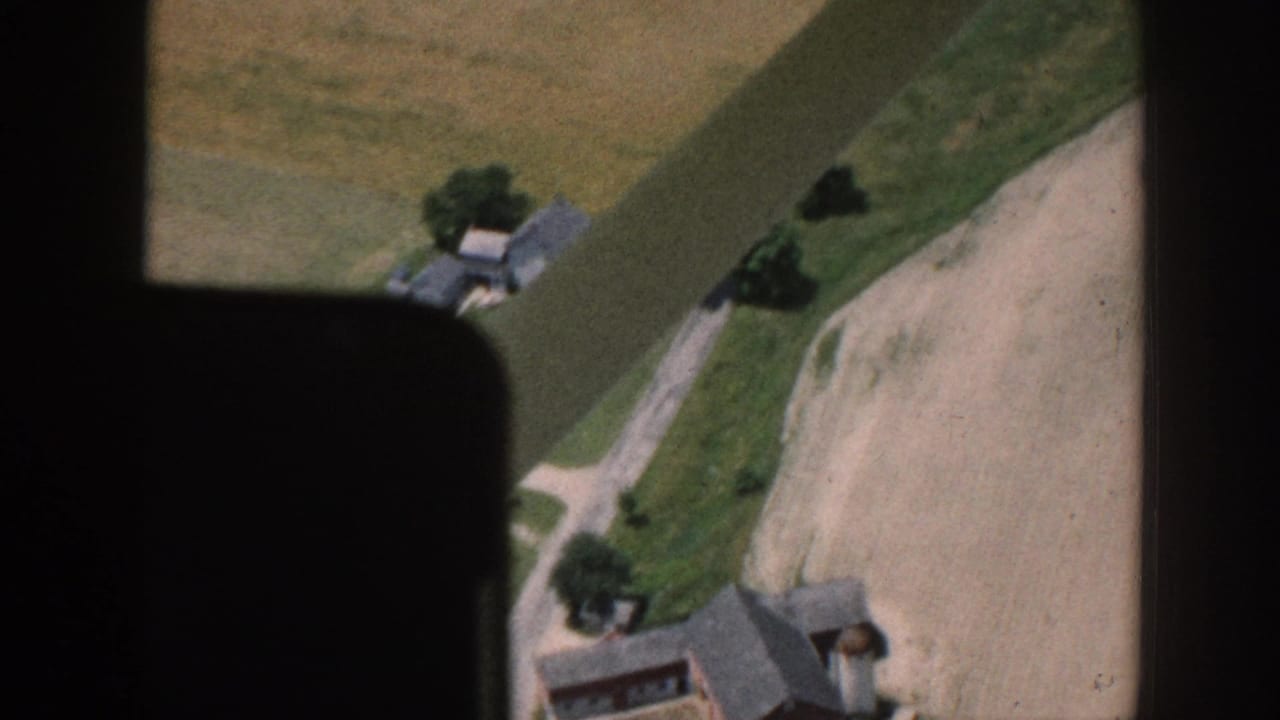 Aerial 8mm view through an aircraft window over what metadata identifies as rural Nebraska, circa 1958, showing farmhouses, a dirt road, and a bare plowed field.