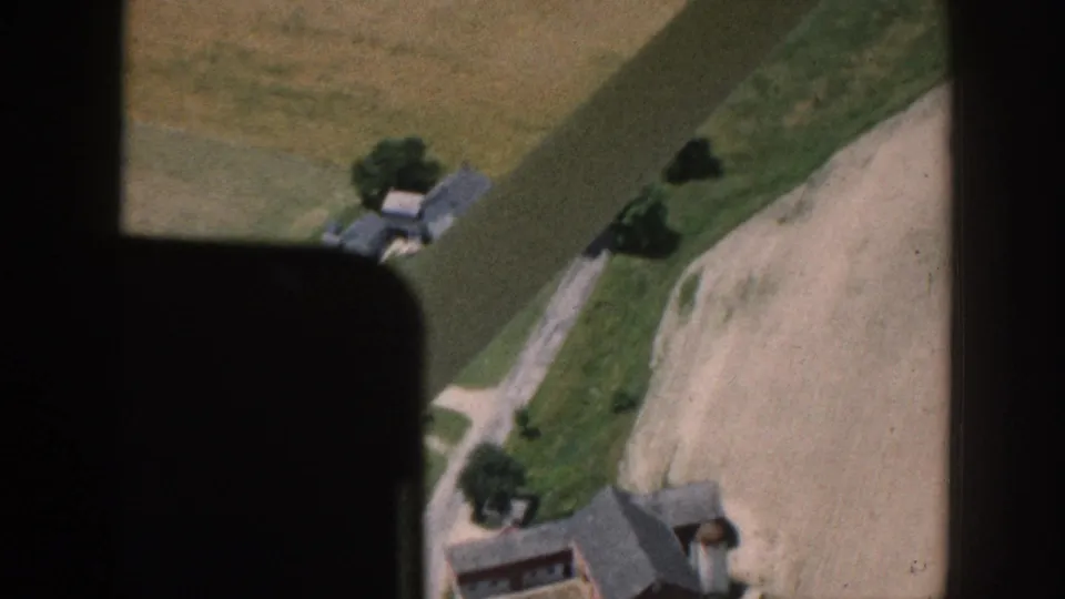 Aerial 8mm view through an aircraft window over what metadata identifies as rural Nebraska, circa 1958, showing farmhouses, a dirt road, and a bare plowed field.