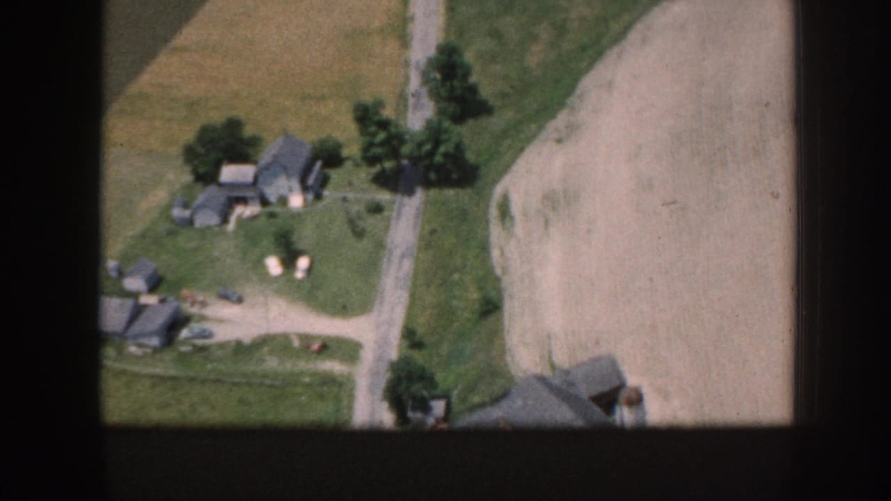 Aerial 8mm view over a rural farmstead in what metadata identifies as Nebraska, 1958, showing outbuildings, a dirt road, plowed field, and apparent small livestock visible in the yard.