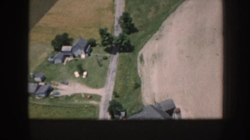 Aerial 8mm view over a rural farmstead in what metadata identifies as Nebraska, 1958, showing outbuildings, a dirt road, plowed field, and apparent small livestock visible in the yard.