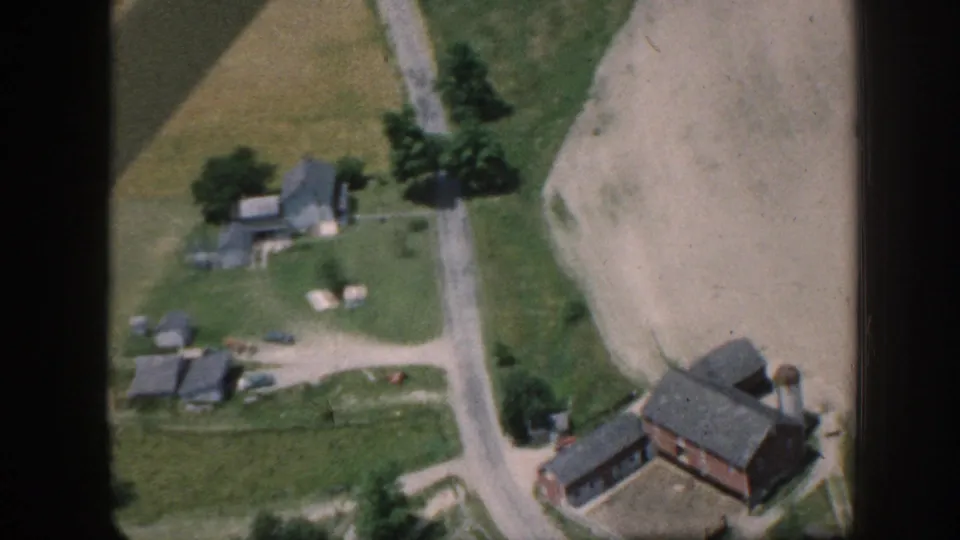 Low aerial 8mm view over a Nebraska farmstead, circa 1958, showing multiple farm buildings, a grain silo or water tower, dirt roads, a parked vehicle, and apparent livestock in the yard.