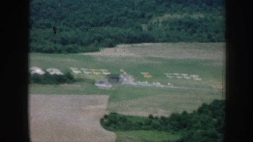 Oblique aerial 8mm view over rural Nebraska, circa 1958, with a dense tree line in the background, green agricultural fields, and what may be farm equipment or structures in the middle distance.