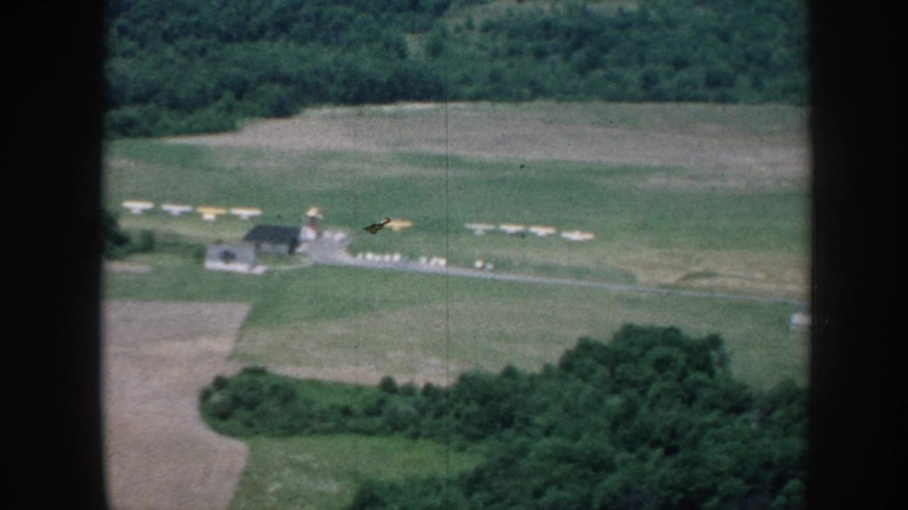 8mm aerial shot circa 1958 looking down on a small Nebraska grass airstrip with parked light aircraft, a single low structure, and a surrounding treeline.