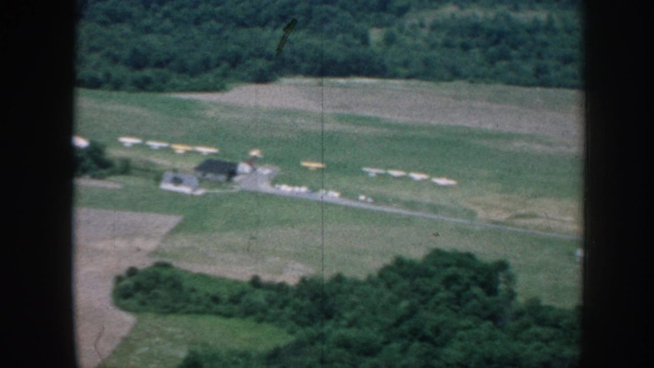 Continued aerial pan circa 1958 over a small Nebraska grass airstrip, showing parked light aircraft, a low structure, and mixed farmland and woodland.