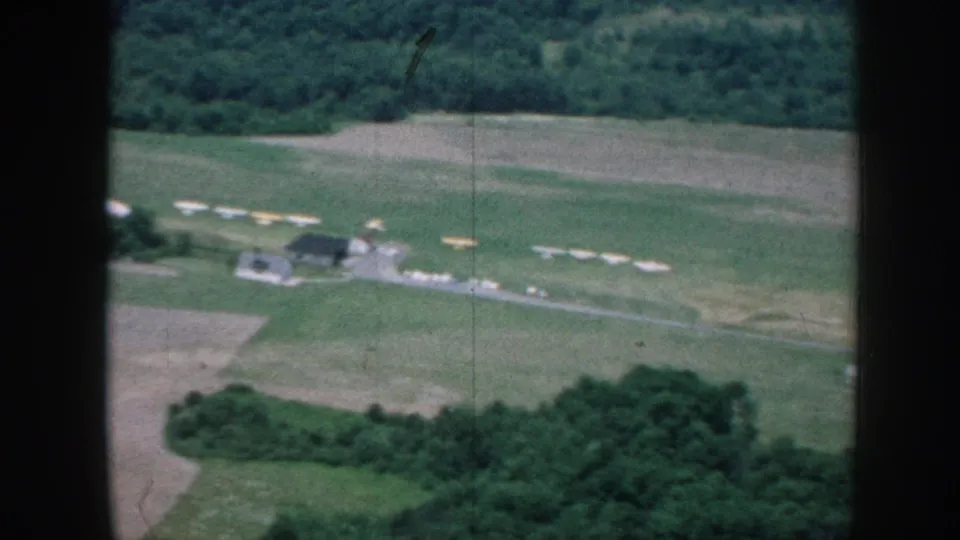 Continued aerial pan circa 1958 over a small Nebraska grass airstrip, showing parked light aircraft, a low structure, and mixed farmland and woodland.