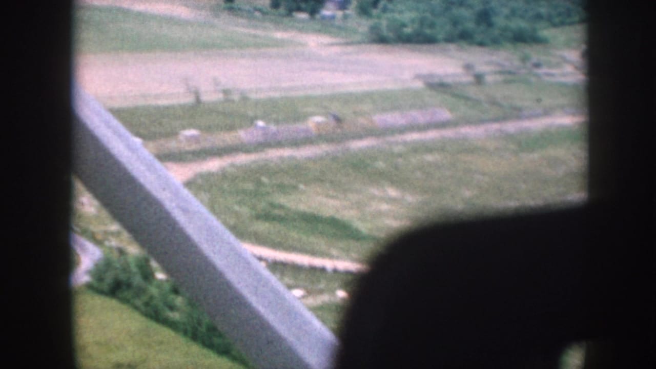 Aerial view through aircraft struts over patchwork farmland with a winding road and small structures.