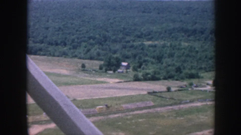 1958 8mm aerial view through light-aircraft struts over a forested hillside, open agricultural fields, and a small farm building below, Nebraska.