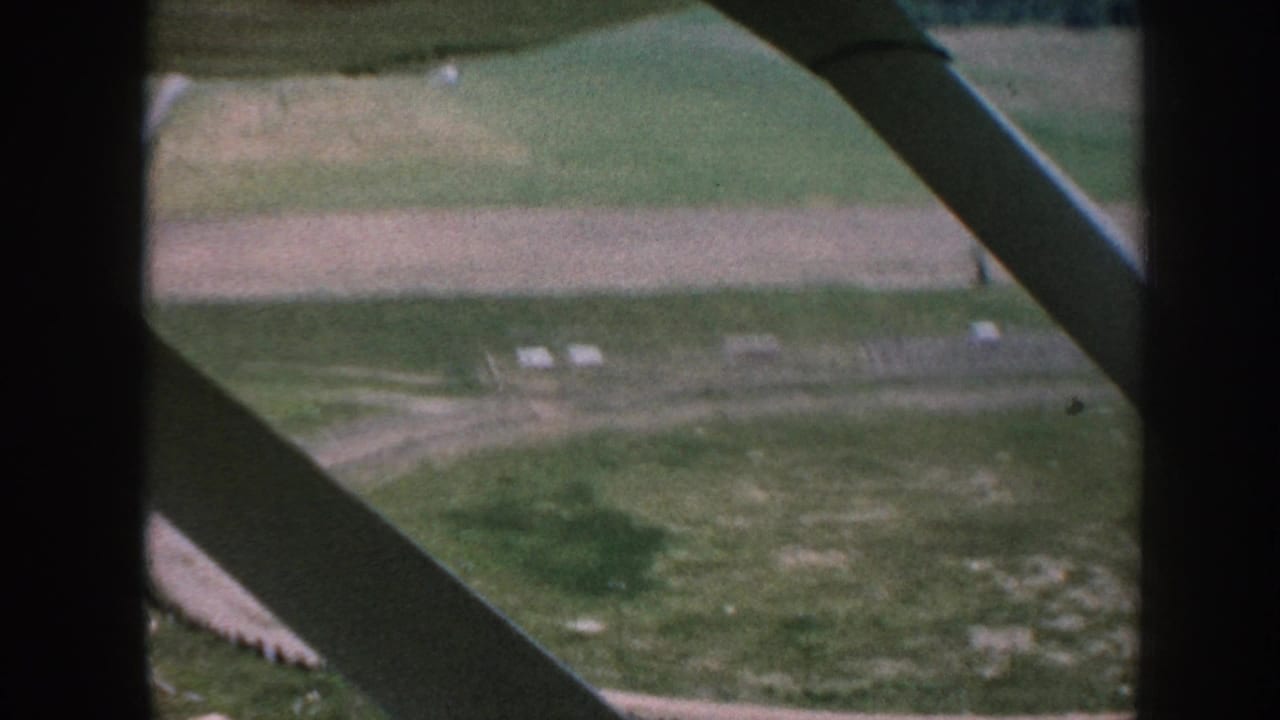 1950s 8mm aerial shot from a small plane interior showing green and plowed farmland below, with a diagonal structural strut and dark window-frame edge visible.