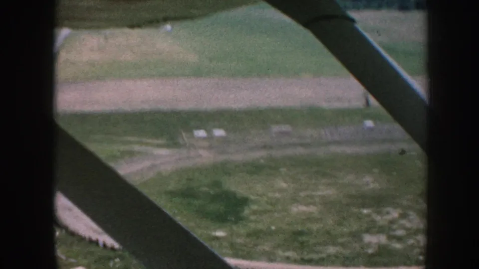 1950s 8mm aerial shot from a small plane interior showing green and plowed farmland below, with a diagonal structural strut and dark window-frame edge visible.