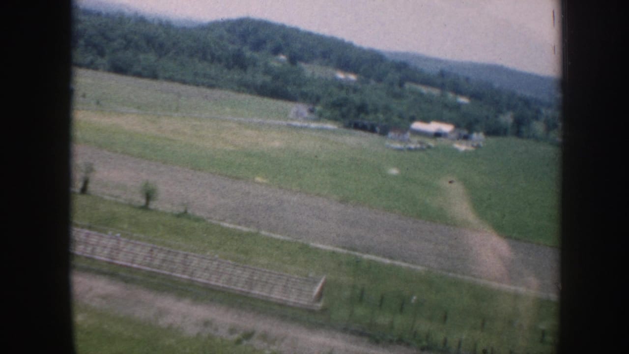 Aerial view of green farmland with a long fence line, white farm outbuildings in mid-distance, and wooded rolling hills on the horizon under a pale hazy sky.