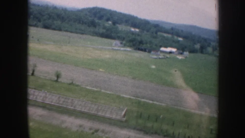1958 8mm aerial footage showing patchwork farmland, a fence line or hedgerow, white farm outbuildings to the right, and wooded rolling hills under a pale hazy sky.