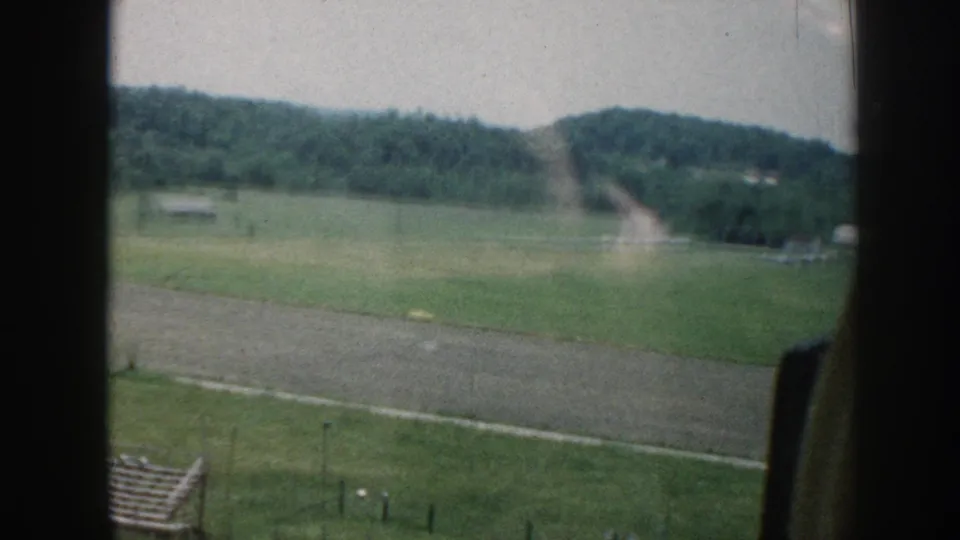 Late-1950s 8mm aerial footage showing green farm fields, a dirt track running through the landscape, wooded rolling hills, fence lines at the bottom, and the aircraft window frame at right.