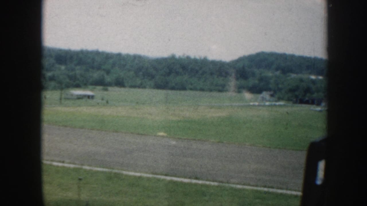 1958 Nebraska countryside filmed through a car window, showing flat green fields, a tilled strip, and distant farmsteads under a hazy sky.