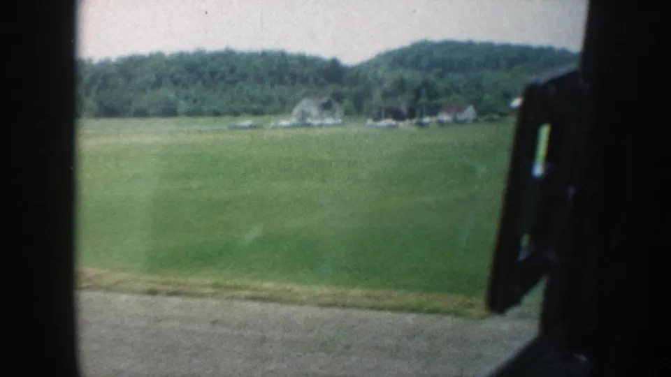 Nebraska summer pasture circa 1958 filmed from a moving car; chrome window latch in foreground, farmstead and forested hills in the distance.