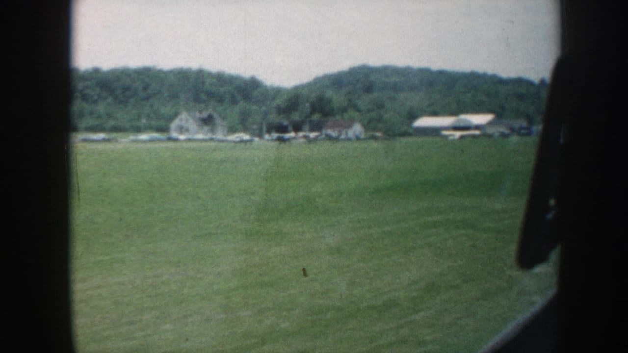 8mm home movie frame, Nebraska c. 1958: rolling green pasture with white clapboard farmhouses and barn structures against a wooded ridge, shot through a car window.