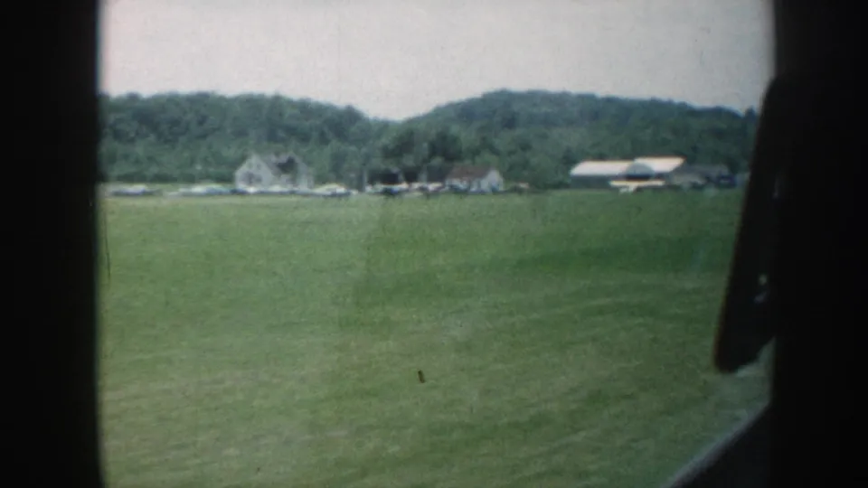 8mm home movie frame, Nebraska c. 1958: rolling green pasture with white clapboard farmhouses and barn structures against a wooded ridge, shot through a car window.