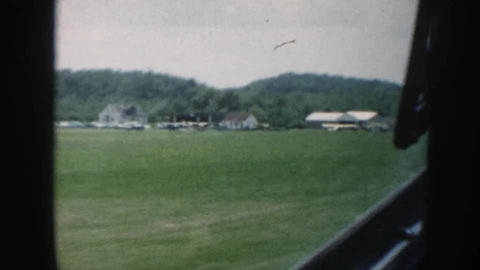 Continued 8mm road-travel frame, Nebraska c. 1958: farmstead cluster and wooded hillside visible as vehicle moves forward, with vehicle door frame at right edge.