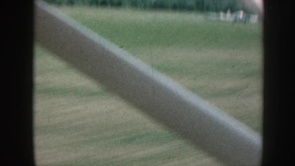 Late-clip 8mm frame, Nebraska c. 1958: vehicle interior trim dominates lower-right foreground, with green pasture and small indistinct distant elements visible at left.