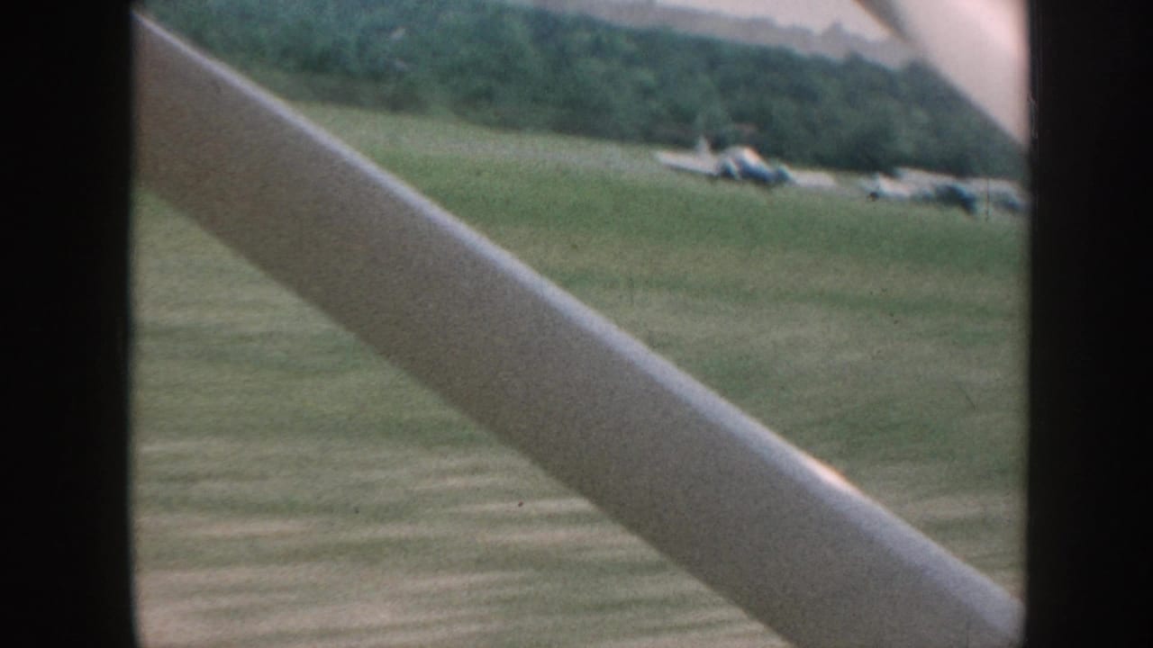 8mm home movie frame, Nebraska 1958: grass airfield seen through a diagonal aircraft strut, parked single-engine planes in the middle distance, forested hills on the horizon.