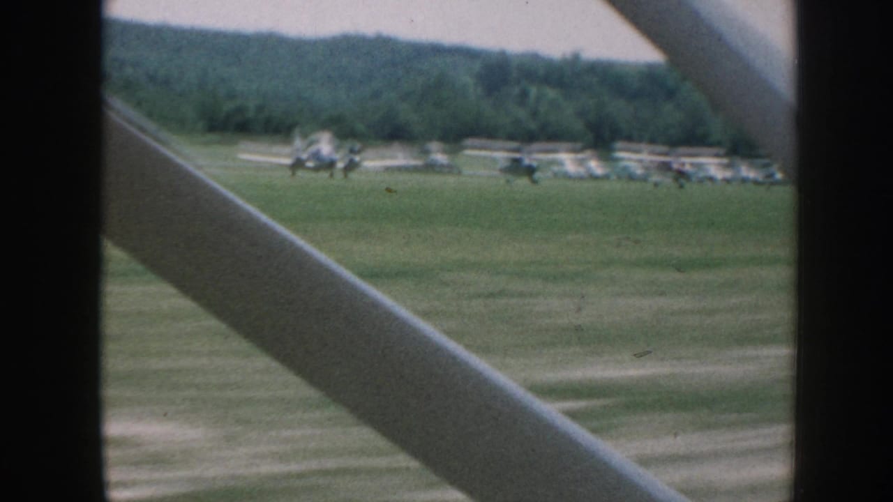 8mm shot from beside an aircraft, Nebraska 1958: multiple small planes parked on a grass airfield with a dense tree-line horizon, diagonal strut crossing the frame.