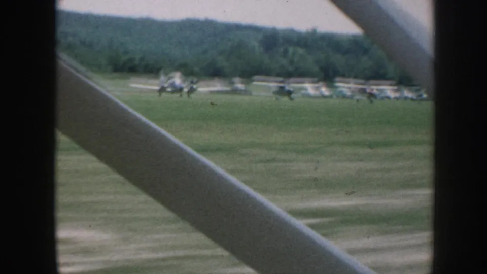 8mm shot from beside an aircraft, Nebraska 1958: multiple small planes parked on a grass airfield with a dense tree-line horizon, diagonal strut crossing the frame.