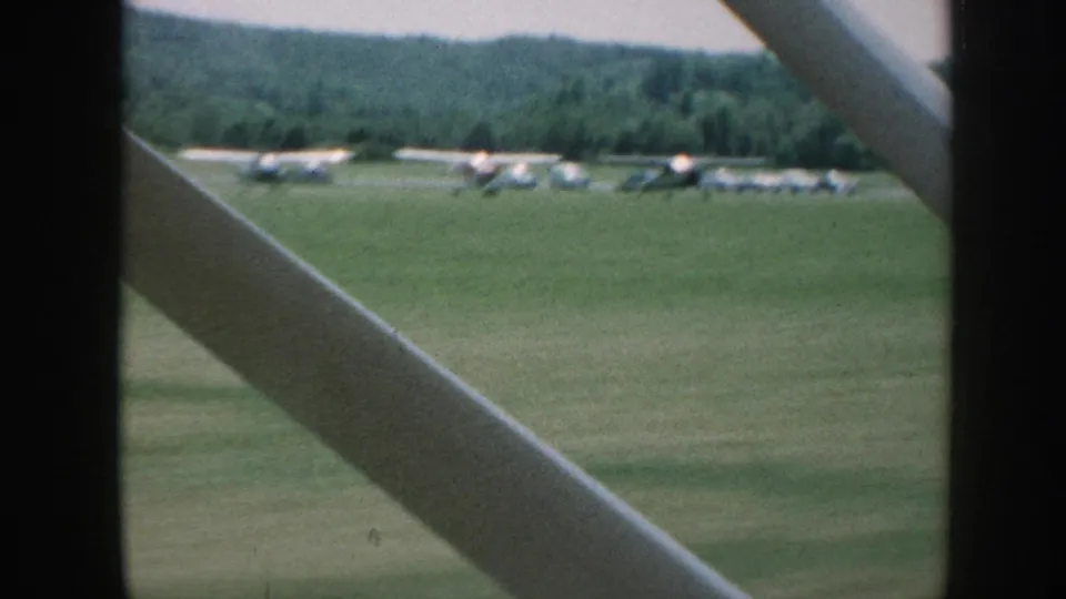8mm frame, Nebraska grass airfield circa 1958: extended row of parked single-engine planes along the field boundary, wooded hills behind, strut at lower-left corner.