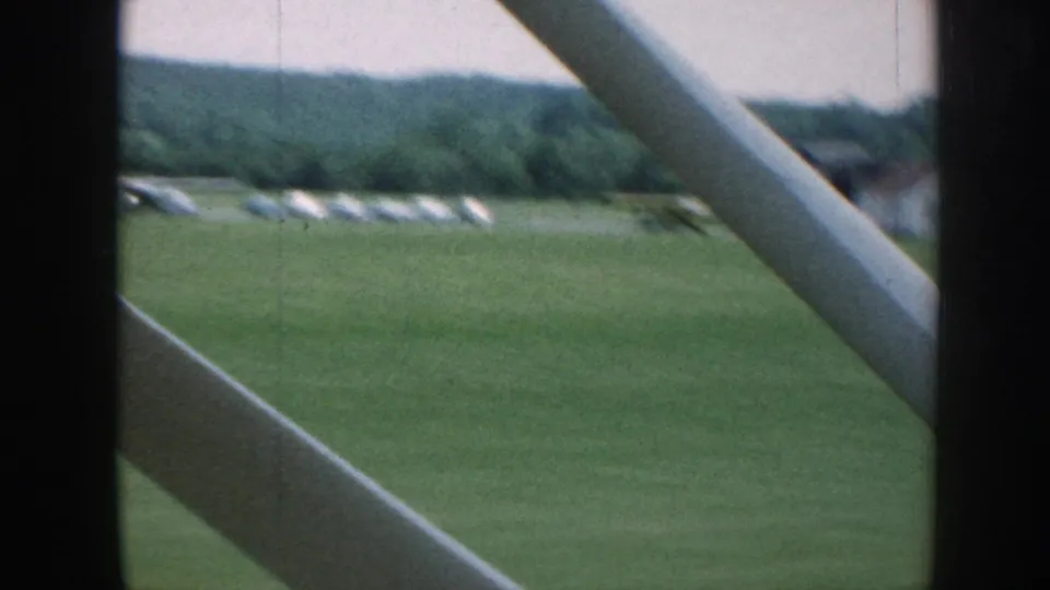 Final 8mm frame, Nebraska airfield circa 1958: row of parked planes with apparent takeoff motion in mid-field, aircraft strut at lower left, wooded horizon.